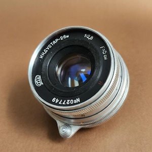 May include: A silver and black camera lens with the text "ИНДУСТАР-26м" and "1:2,8 F=5 cm" printed on the lens. The lens has a ridged focusing ring and the serial number "N027749".