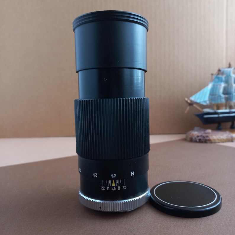 Op de afbeelding: Een zwarte cameralens met een geribbelde grip en een zilveren ring aan de basis. De lens heeft markeringen voor f-stops en een losse zwarte lensdop. Een miniatuur zeilboot is zichtbaar op de achtergrond.