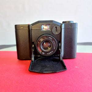 Puede incluir: Una cámara vintage negra con una lente que dice "MC KOPCAP 2.8/35". La cámara tiene un flash y un pequeño visor. La tapa de la lente está abierta, revelando la lente. La cámara está sobre una superficie roja.