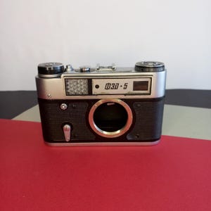 以下が含まれることがあります： 黒いボディとシルバーのトップを備えた、ヴィンテージのФЭД-5カメラ。 カメラには大きなレンズ開口部と「ФЭД-5」の文字が入った小さな窓があります。 カメラは赤い面に置かれています。