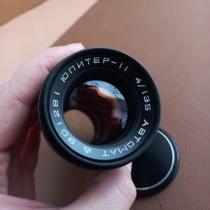 Op de afbeelding: Een zwarte cameralens met de tekst "ЮПИТЕР-11" en "4/135" op de rand. De lens is gedeeltelijk bedekt door een zwarte lensdop. De lens wordt in een hand gehouden.