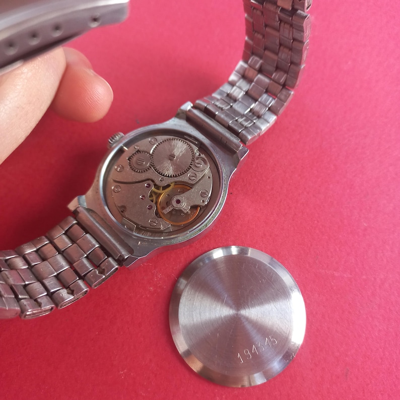 May include: A silver-toned wristwatch with a metal band, showcasing the intricate internal mechanisms. The watch's back is detached, revealing gears and springs. The watch's back cover is also visible, with the number 194545.