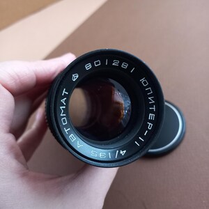 Op de afbeelding: Zwarte cameralens met de tekst "801281", "Jupiter-11" en "A/135". De lens wordt in een hand gehouden en heeft een zwarte lensdop.