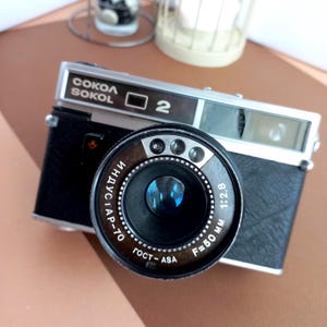 May include: A vintage Sokol 2 rangefinder camera with a black textured body and silver metal top plate. The lens is marked Industar-70, F=50 mm, 1:2.8, and GOST-ASA. The front features a prominent circular lens with three small light sensors.