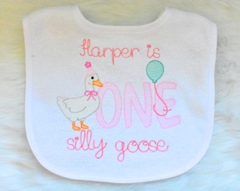 May include: A white baby bib featuring embroidered text that reads Harper is ONE silly goose. The design includes a white goose wearing a pink party hat and bow, alongside a light green balloon and large pink letters spelling ONE.
