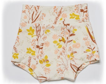May include: High-waisted shorts with a floral pattern. The shorts feature a cream-colored background with a repeating pattern of yellow, peach, and brown flowers and foliage. The shorts have a folded hem and a wide waistband.