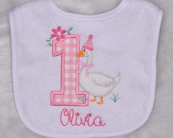 May include: White baby bib with a pink gingham number one and a goose wearing a party hat. The bib has the name "Olivia" embroidered in pink script. The goose has a pink bow and is standing on green grass.