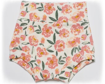 May include: High-waisted baby shorts with a floral pattern. The shorts feature a cream-colored background with a repeating pattern of pink and yellow flowers and green leaves. The fabric is soft and the shorts are designed for comfort.