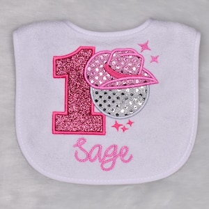 May include: White baby bib with a pink glitter number 1 and a pink cowboy hat design. The bib has the name "Sage" embroidered in pink script. The design includes pink stars.