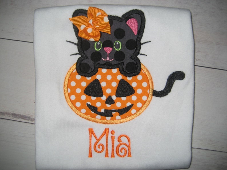 May include: White shirt with an embroidered Halloween design. A black cat with polka dots sits atop an orange pumpkin with a jack-o'-lantern face. The name "Mia" is embroidered in orange below the pumpkin.