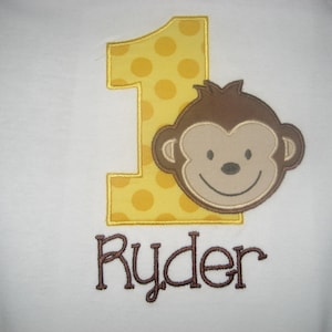 May include: White onesie with a yellow polka-dot number one and a brown monkey face applique. The name "Ryder" is embroidered in brown script below the number and monkey. A cute outfit for a baby's first birthday.