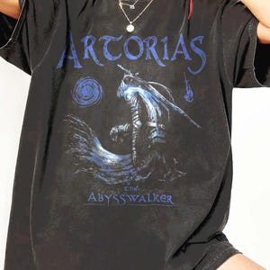 Op de afbeelding: Zwart T-shirt met de blauwe tekst "ARTORIAS" en een blauw-witte illustratie van een krijger. De tekst "the ABYSSWALKER" staat eronder. Het T-shirt heeft korte mouwen.