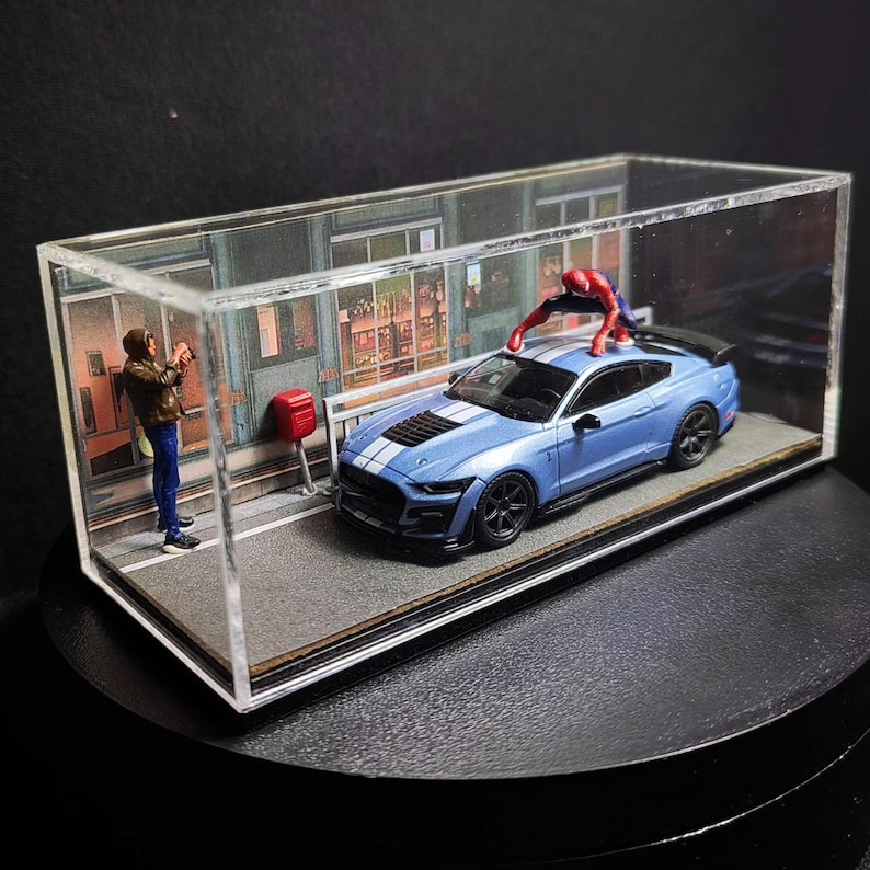 Może przedstawiać: Niebieski model samochodu Ford Mustang Shelby GT500 w przezroczystej gablocie z figurką Spidermana w kolorze czerwonym i niebieskim na dachu samochodu. Osoba w brązowej kurtce stoi po lewej stronie samochodu, trzymając aparat.
