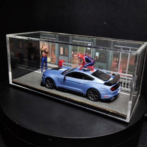 Może przedstawiać: Niebieski Ford Mustang ze spojlerem w kolorze czarnym i figurką Spider-Mana w kolorze czerwonym i niebieskim na dachu. Samoch&oacute;d znajduje się w szklanej gablocie z tłem przedstawiającym ulicę miasta.