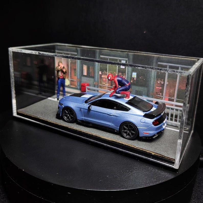 Może przedstawiać: Niebieski Ford Mustang ze spojlerem w kolorze czarnym i figurką Spider-Mana w kolorze czerwonym i niebieskim na dachu. Samoch&oacute;d znajduje się w szklanej gablocie z tłem przedstawiającym ulicę miasta.