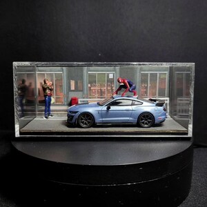 Może przedstawiać: Niebieski model samochodu Ford Mustang Shelby GT500 w szklanej gablocie z figurką Spidermana na dachu samochodu. W tle stoi osoba w niebieskiej koszuli i dżinsach.