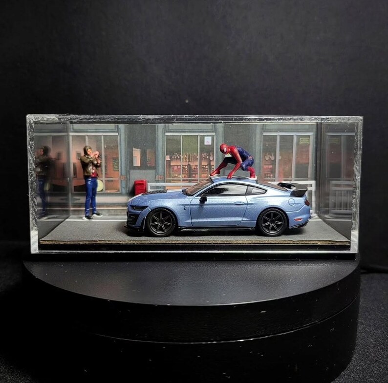 Może przedstawiać: Niebieski model samochodu Ford Mustang Shelby GT500 w szklanej gablocie z figurką Spidermana na dachu samochodu. W tle stoi osoba w niebieskiej koszuli i dżinsach.