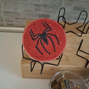 Op de afbeelding: Een ronde, rode onderzetter met een zwart spinnenmotief. De spin is gecentreerd op de onderzetter en is gemaakt van zwarte strass-steentjes. De onderzetter staat op een zwarte metalen standaard.