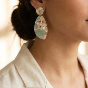 May include: A pair of abstract-patterned earrings. The earrings feature a small, round stud and a larger, teardrop-shaped pendant. The color palette includes shades of green, beige, and brown. The earrings are a unique accessory.