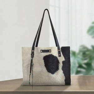 May include: A tote bag crafted from cowhide featuring a black and white spotted pattern. The bag includes two long black leather shoulder straps, a rectangular leather patch on the front, and a decorative leather tassel with small turquoise beads.