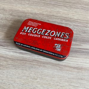 Puede incluir: Una lata rectangular roja con la palabra "MEGGEZONES" en letras blancas grandes. La lata también tiene el texto "FOR COUGHS COLDS CATARRH" y "MADE IN LONDON, ENGLAND, BY MEGGESON & CO., LTD."