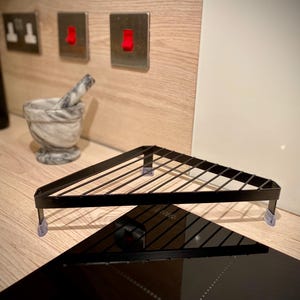 May include: Black metal triangular shelf with a wire grid design. The shelf is mounted on a worktop with clear adhesive pads.