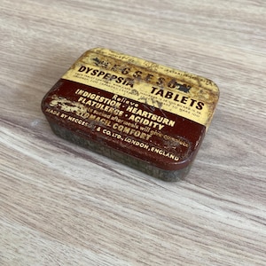 Puede incluir: Una lata rectangular vintage de tabletas Eggeson Dyspepsia, con una combinación de colores marrón y dorado descoloridos. La etiqueta de la lata enumera los ingredientes y el texto "Relieve Indigestion, Heartburn, Flatulence, Acidity." Fabricado por Meggeson & Co. Ltd., Londres, Inglaterra.