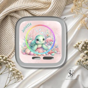 Peut inclure: Un objet décoratif carré gris clair avec une tortue de mer de dessin animé à l'intérieur d'un cercle aux couleurs de l'arc-en-ciel. La tortue est entourée de coraux, de coquillages et du nom "Juliette". L'objet comporte deux petites fentes horizontales en bas.