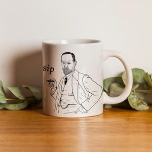 May include: White ceramic mug with a black and white illustration of a man holding a cigar. The word "sip" is printed on the side. The mug has a curved handle and is on a wooden surface.