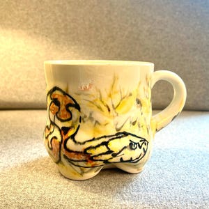 Pode incluir: Uma caneca de cerâmica branca com um desenho de cobra amarela, laranja e preta. A caneca tem um corpo arredondado e uma alça.