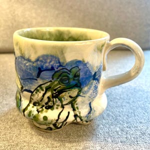 May include: A white ceramic mug with a green, blue, and white frog design. The mug has a rounded bottom and a handle.