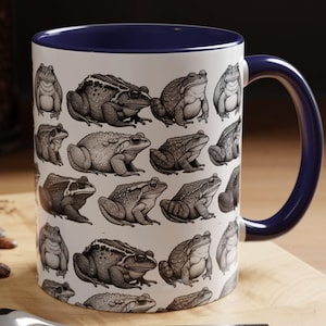 May include: White ceramic mug with a blue rim and handle. The mug features a repeating pattern of black and white illustrations of frogs.