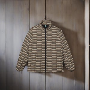 Könnte beinhalten: Eine gesteppte Jacke mit einem geometrischen Muster in Braun-, Beige- und Grautönen. Die Jacke hat einen durchgehenden schwarzen Reißverschluss und einen Stehkragen. Sie hängt an einem schwarzen Kleiderbügel vor einem Holzgrund.