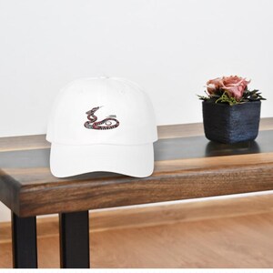 May include: A white baseball cap with a snake design in red, black, and brown. The cap is sitting on a wooden table next to a small potted plant with pink flowers.