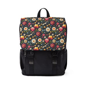 Puede incluir: Una mochila negra con solapa con un colorido estampado floral. El estampado incluye flores rojas, amarillas y blancas, estrellas y libélulas sobre un fondo azul oscuro. La mochila tiene correas y hebillas negras.
