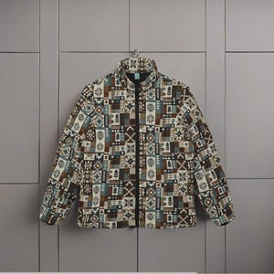 Op de afbeelding: Een jas met een hoge kraag en een lange ritssluiting, met een patchwork-ontwerp. Het patroon bevat bruine, beige, blauwe en zwarte vierkanten. De jas hangt aan een haak aan een grijze muur.