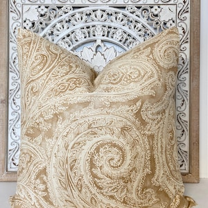 May include: A beige and white paisley patterned throw pillow with a decorative fringe. The pillow is in front of a white wooden decorative wall panel.