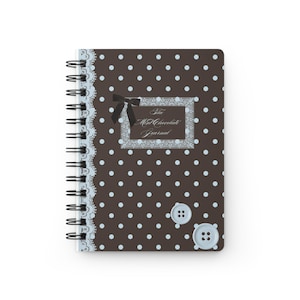 Puede incluir: Un cuaderno de espiral con cubierta marrón oscuro con lunares azul claro, una franja vertical de encaje azul claro, un lazo negro y dos botones azul claro. Una etiqueta rectangular central muestra el texto The Mint Chocolate Journal.