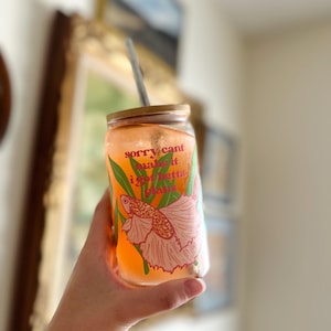 Puede incluir: Un vaso de vidrio transparente con tapa de bambú y pajita. El vaso contiene un líquido naranja y presenta un diseño de pez betta rosa y naranja con el texto "sorry cant make it i got betta plans".