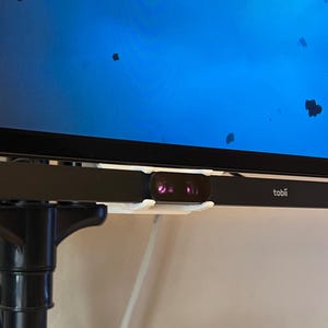 May include: A black webcam with a white mount attached to the bottom of a computer monitor. The webcam has a small red light on the front. The text "tobii" is visible on the webcam.