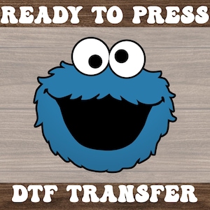 May include: A blue cartoon character with large white eyes and a wide open black mouth. The character is surrounded by blue fur. The words "READY TO PRESS" and "DTF TRANSFER" are printed above and below the character in white.