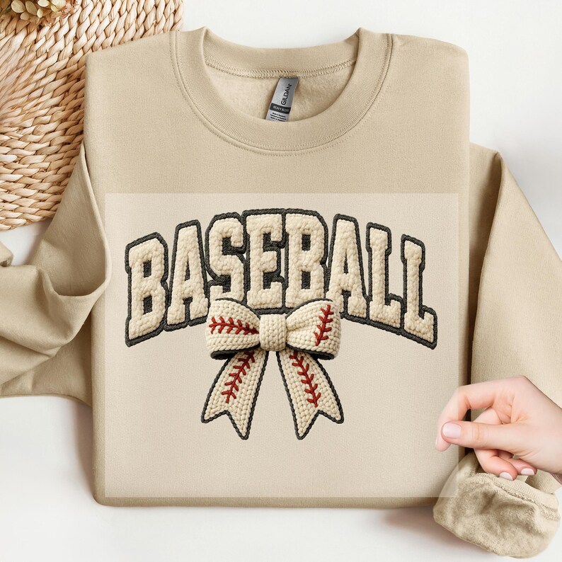May include: A beige sweatshirt with the word "BASEBALL" in a cream-colored, embroidered font. Below the word is a cream-colored bow with red stitching, resembling a baseball. The sweatshirt is a neutral color.