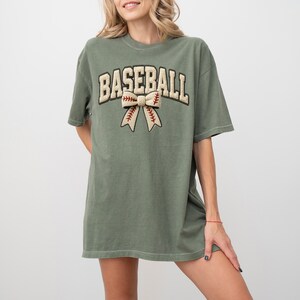 May include: An olive green t-shirt with the word "BASEBALL" in a cream-colored, collegiate-style font. Below the word is a bow with baseball stitching details. The shirt has short sleeves and a relaxed fit.