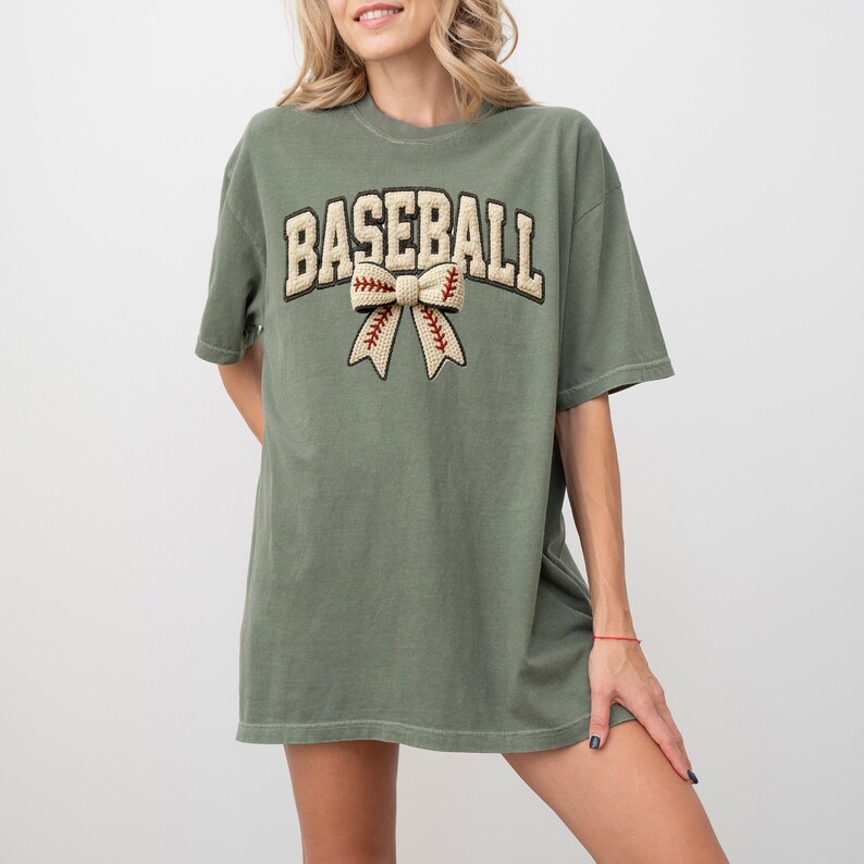 May include: An olive green t-shirt with the word "BASEBALL" in a cream-colored, collegiate-style font. Below the word is a bow with baseball stitching details. The shirt has short sleeves and a relaxed fit.
