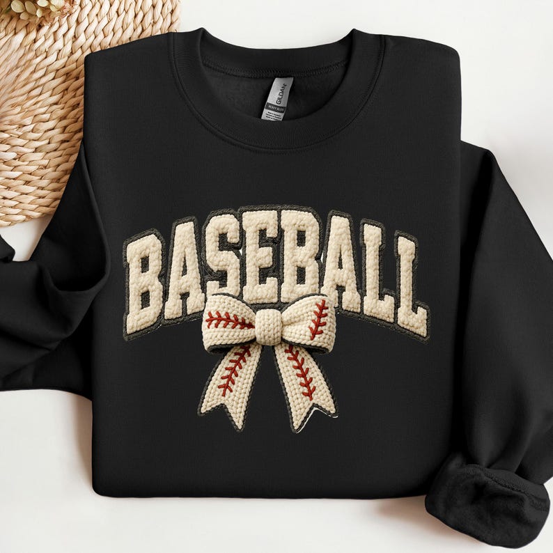May include: Black sweatshirt with the word "BASEBALL" in a cream-colored, textured font. A cream-colored bow with red stitching, resembling a baseball, is centered below the word. The sweatshirt has a crew neck and long sleeves.