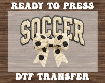 May include: A cream-colored "SOCCER" design with a black outline and a bow featuring black soccer ball patterns. The words "READY TO PRESS" and "DTF TRANSFER" are displayed in a bold, rounded font.