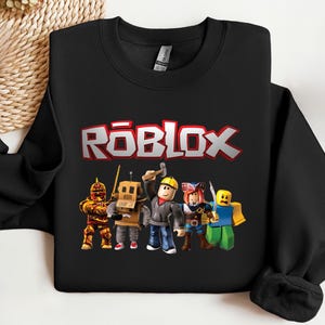 May include: Black sweatshirt featuring the word "ROBLOX" in red and white block letters. Below the text are five cartoon characters in various outfits, including a knight, robot, and construction worker.