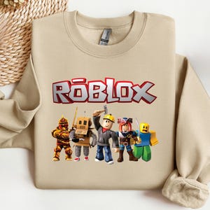 May include: A beige sweatshirt featuring the word "ROBLOX" in red and white block letters. Below the text are cartoon figures of various characters. The sweatshirt is made of a soft material.