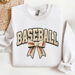 May include: White sweatshirt with the word "BASEBALL" in a beige, textured font. A beige bow with red stitching, resembling a baseball, is centered below the word. The sweatshirt has a crew neck and long sleeves.