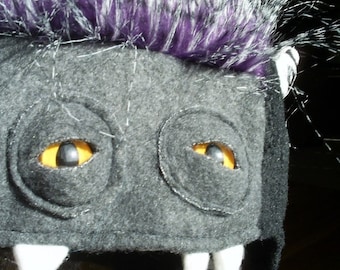 May include: A black and gray plush hat with a purple and white mohawk. The hat has two yellow eyes and white teeth.